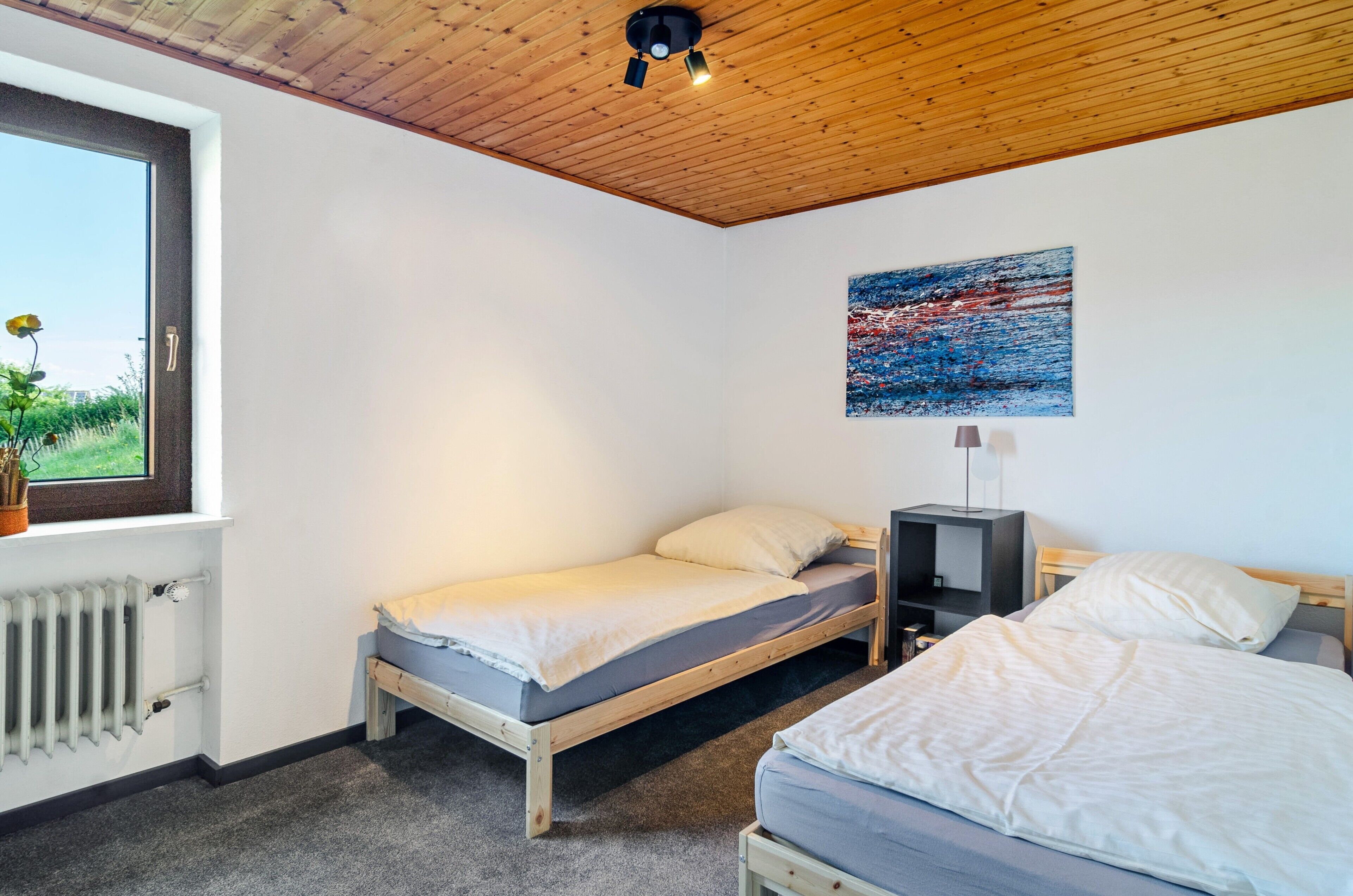 A bedroom with a wooden ceiling, a window with a view, a bed, a bedside table, and a radiator.