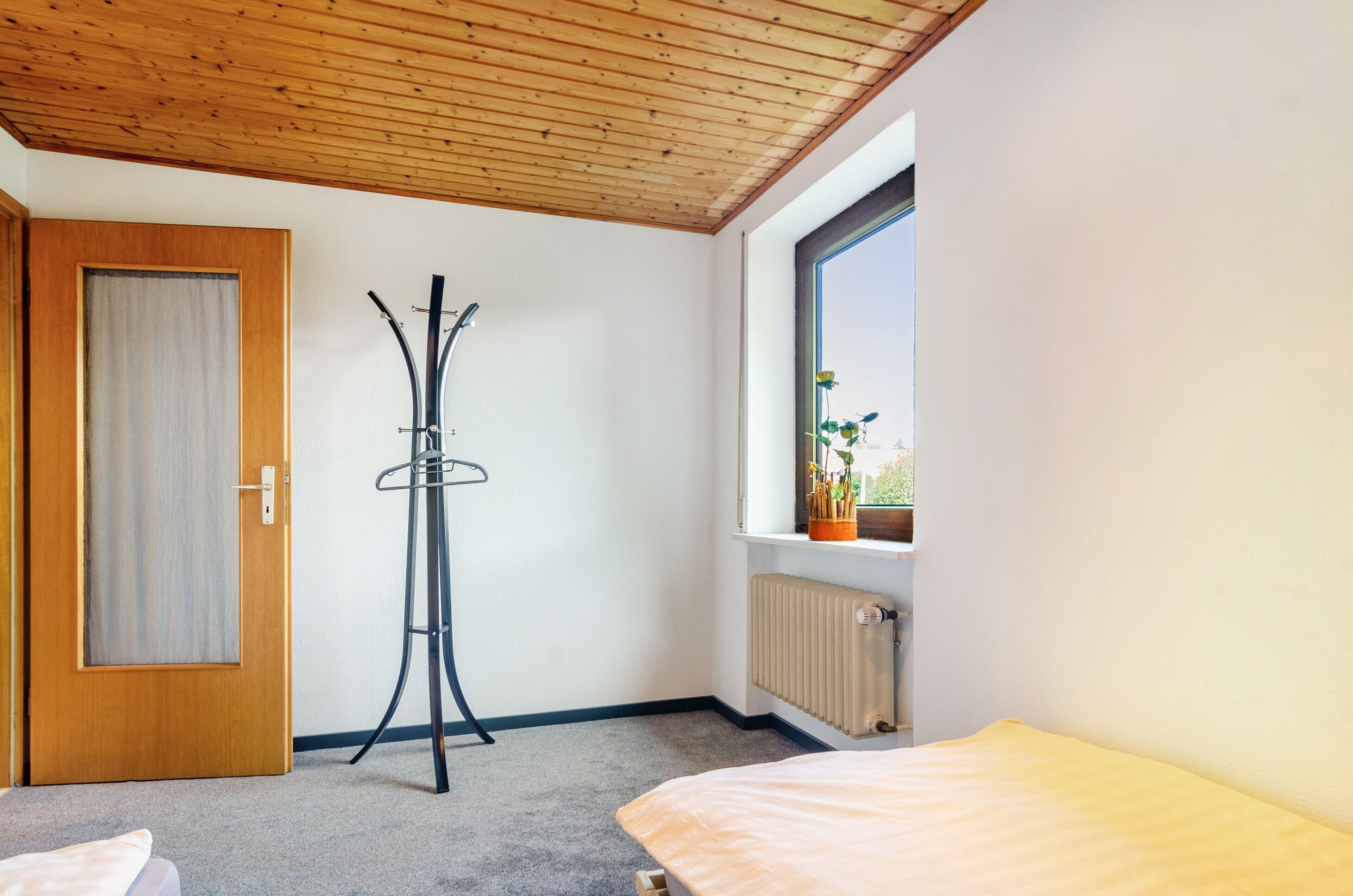 A room with a wooden ceiling, a radiator, a window with a view, a door, and a bed.