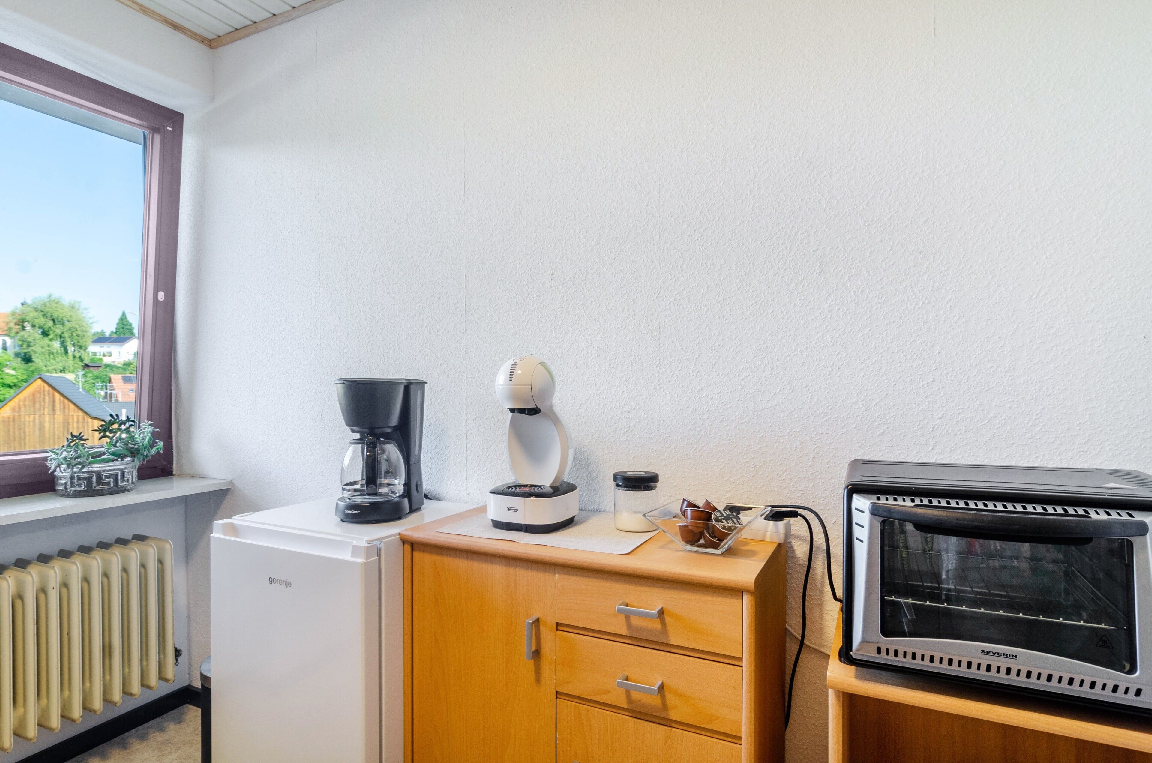 A kitchenette with a coffee maker, a toaster, a small refrigerator, and a window with a view of houses and trees.