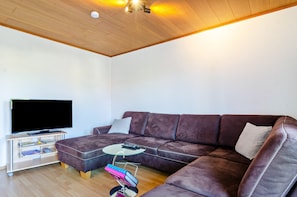 Un salon avec un canapé en cuir marron, une table basse en verre, un téléviseur à écran plat posé sur un meuble et un ventilateur de plafond.