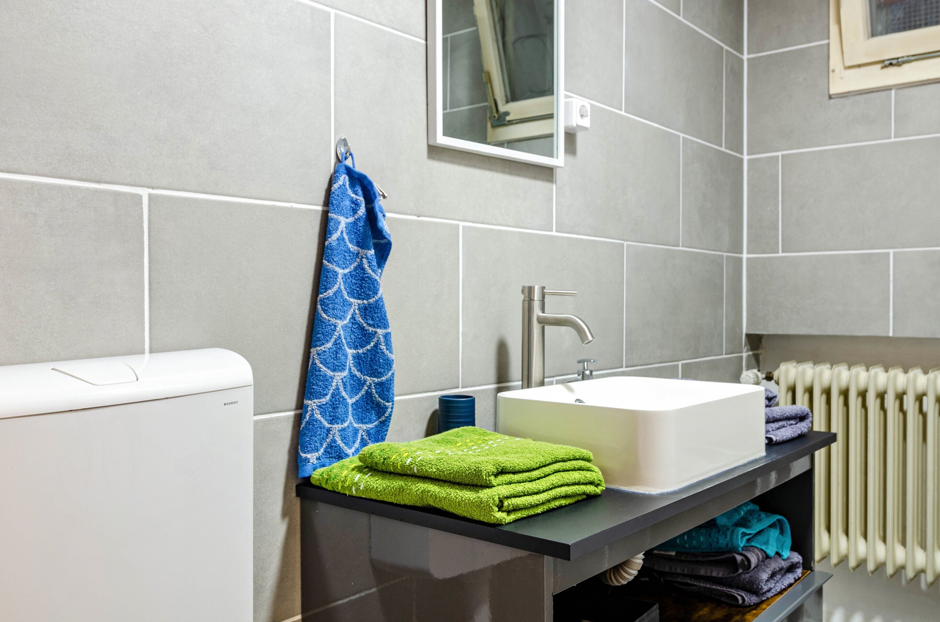A bathroom with grey tiles, a white sink, a blue towel, and a green towel on a black shelf.