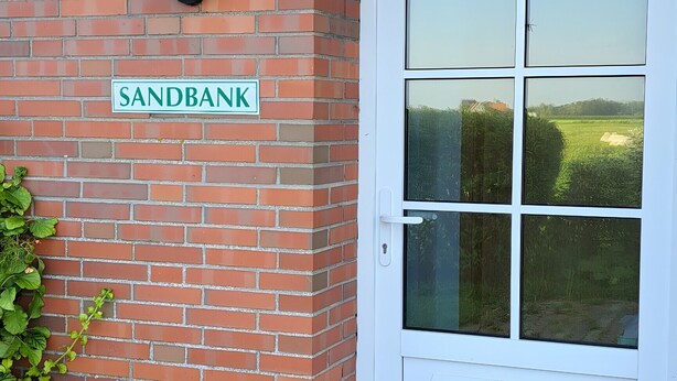 Un bâtiment en briques avec une porte blanche et une enseigne indiquant « SANDBANK ».