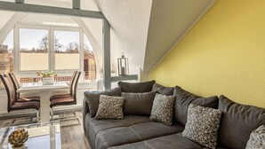 Ein Wohnzimmer mit grauem Sofa, gemusterten Kissen, einem Glastisch und einem Essbereich mit weißem Tisch und Stühlen.