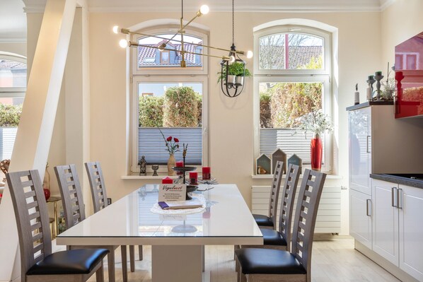 Un espace repas avec une table en verre, des chaises en bois, un luminaire suspendu et des fenêtres donnant sur un paysage verdoyant.