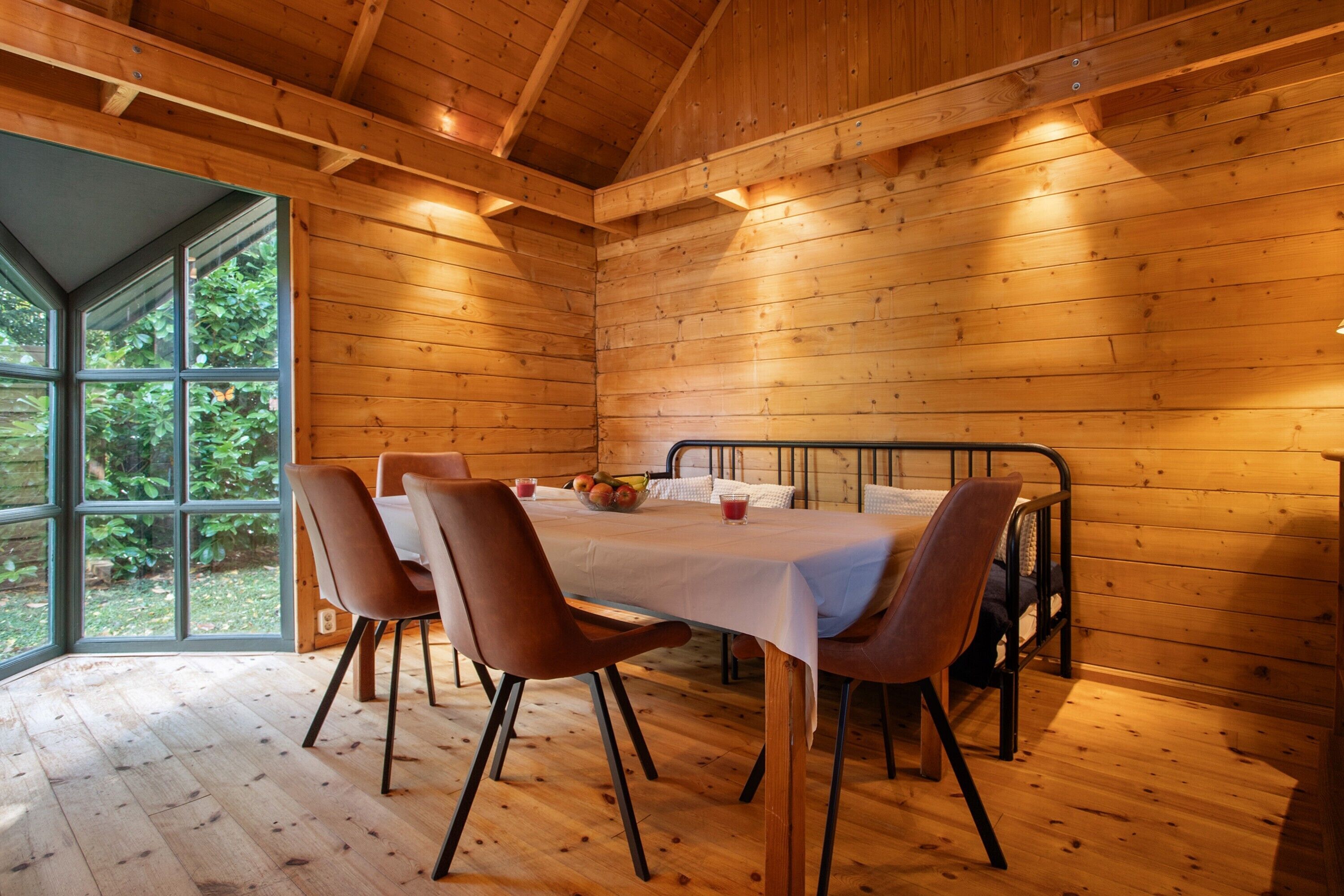 Interior d'una cabana de fusta amb taula de menjador, cadires i un llit amb estructura metàl·lica.