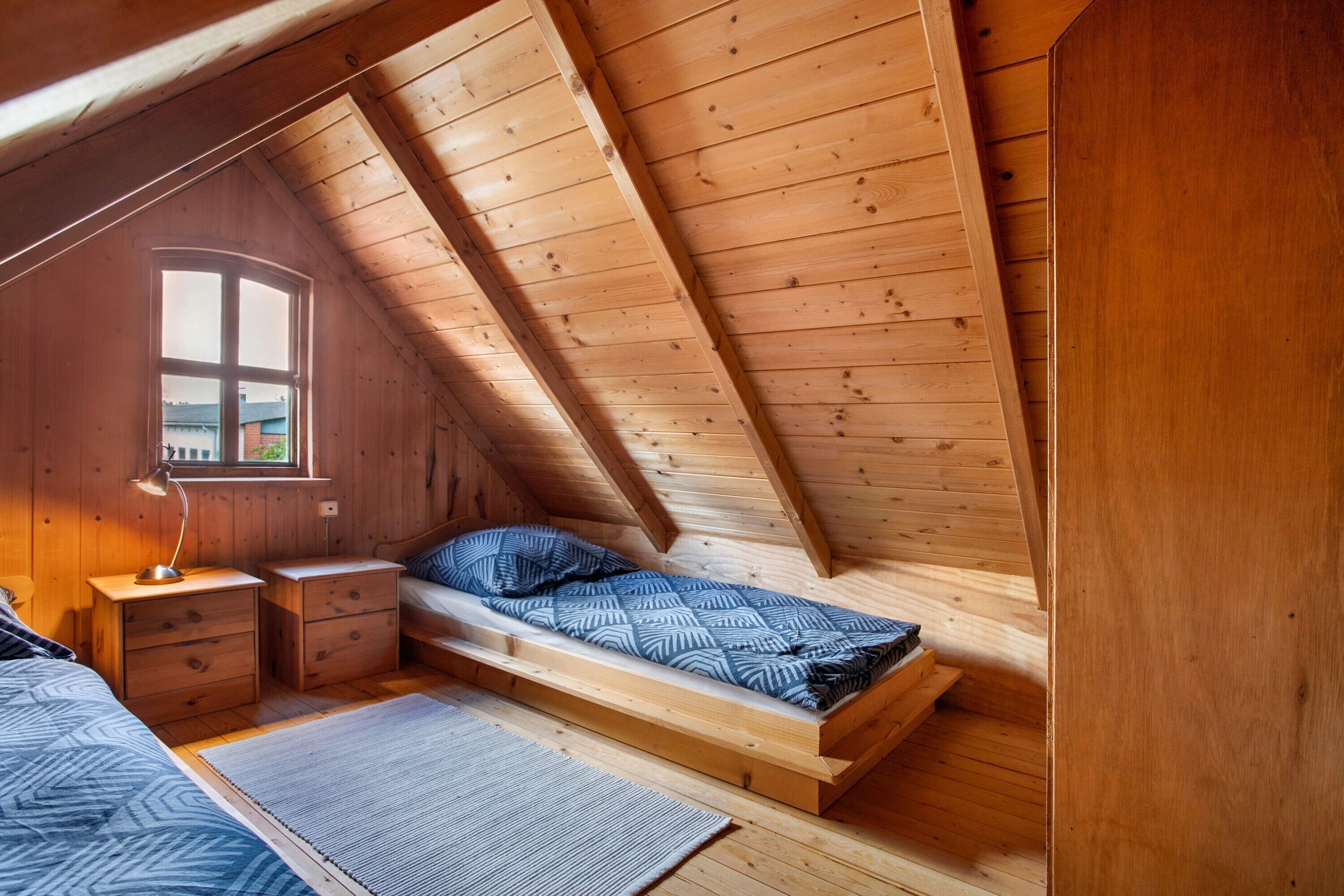 A small, wooden attic bedroom with a single bed, a nightstand, a desk, and a window.