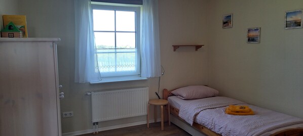 A small bedroom with a single bed, a radiator, a window with curtains, a small table, and a shelf.