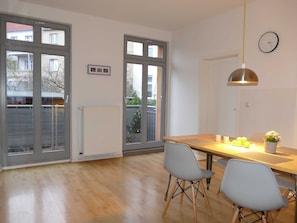 A dining area with a wooden table, two chairs, a pendant light, and a clock on the wall.