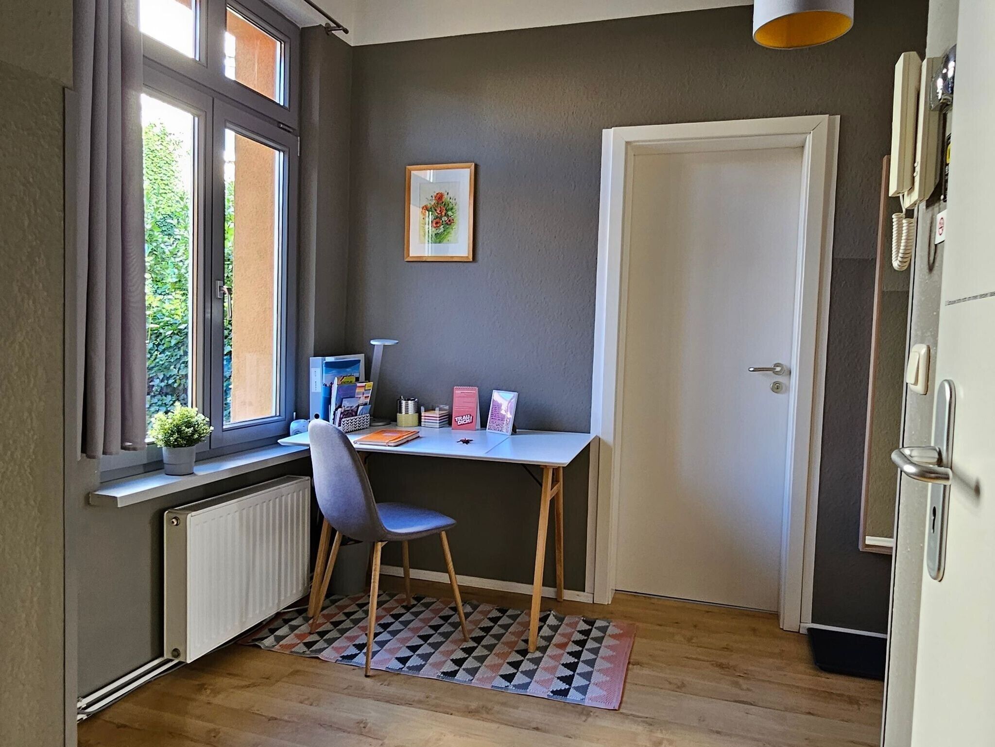 A small room with a desk, chair, radiator, window, and a door.