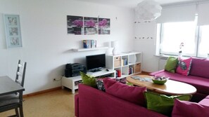 A living room with a TV, bookshelves, a sofa, and a window. - Apartment 'Ferienwohnung Flut' with Shared Garden, Balcony and Wi-Fi (Pellworm)