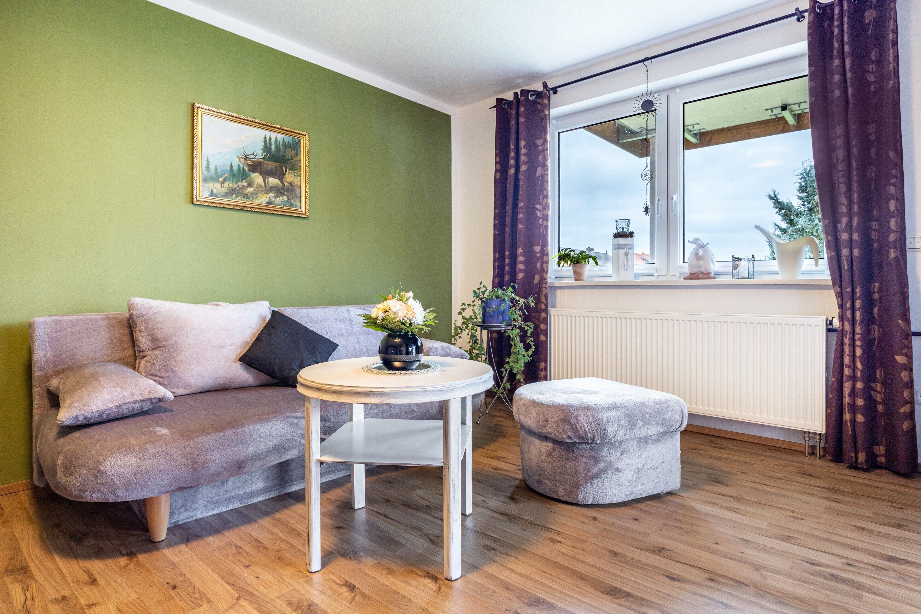 Ein Wohnzimmer mit einer Couch, einem Hocker, einem runden Tisch und einem Fenster mit Vorhängen.