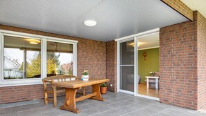 Eine überdachte Terrasse mit einem Holztisch und -stühlen, einem Blumentopf und einer Glasschiebetür, die zu einem Innenraum führt.