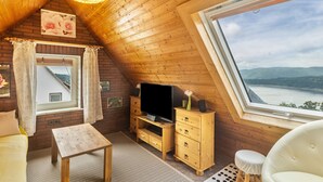 A cozy room with a wooden ceiling, a television on a wooden stand, a window with a view of the landscape, and a small table.