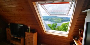 Une pièce avec un plafond en bois, un téléviseur posé sur une commode en bois et un puits de lumière donnant sur un lac et des collines.