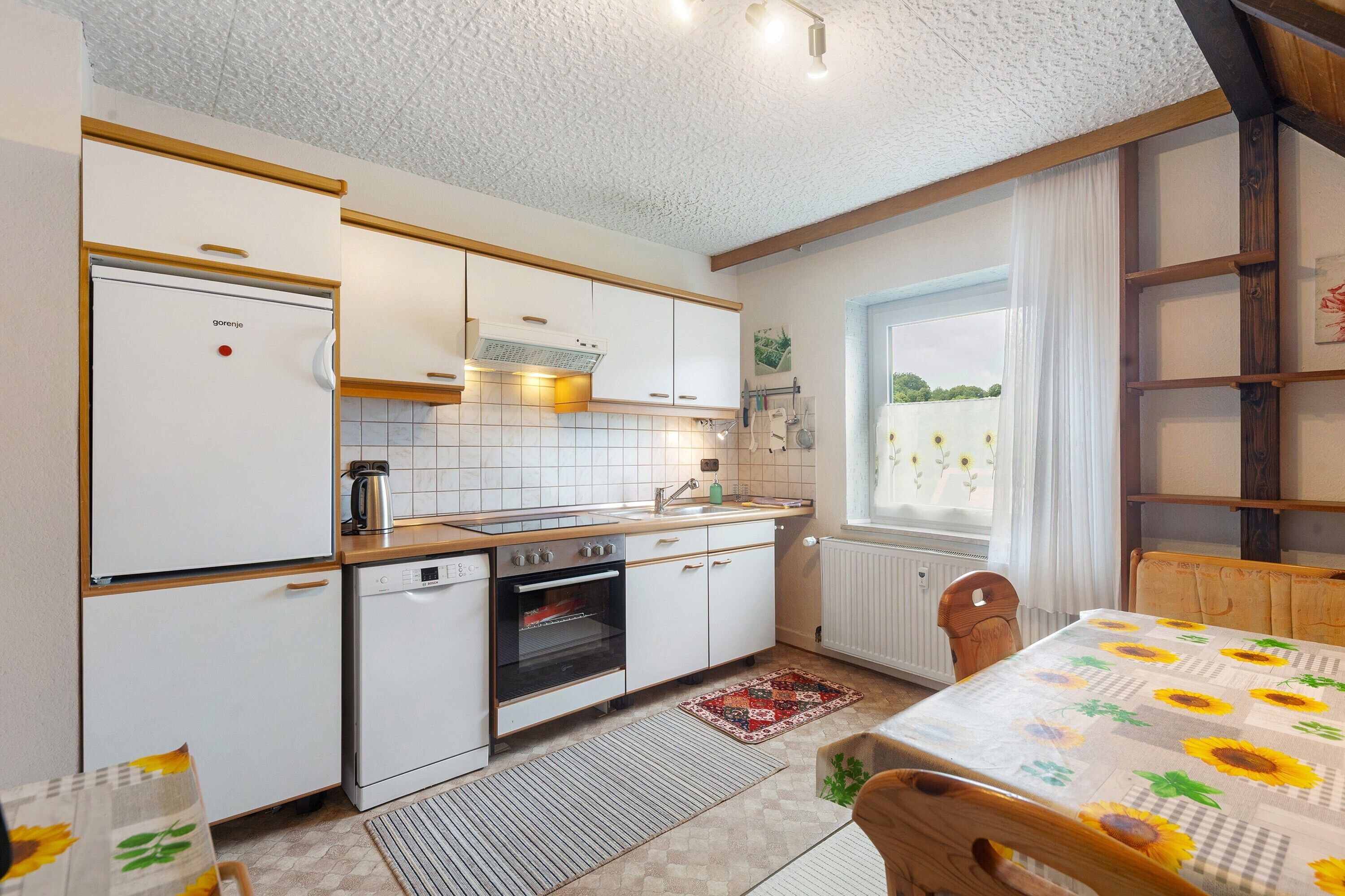 A compact kitchen with white cabinets, a built-in oven, a coffee maker, and a window with a view of greenery.