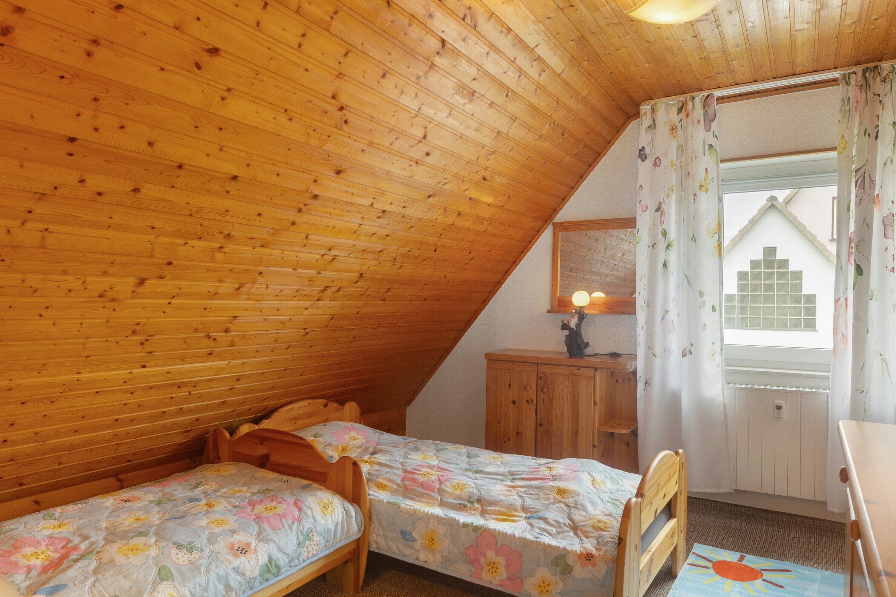 A room with wooden sloped ceilings, two wooden beds with floral bedding, a wooden cabinet, and a window with curtains.