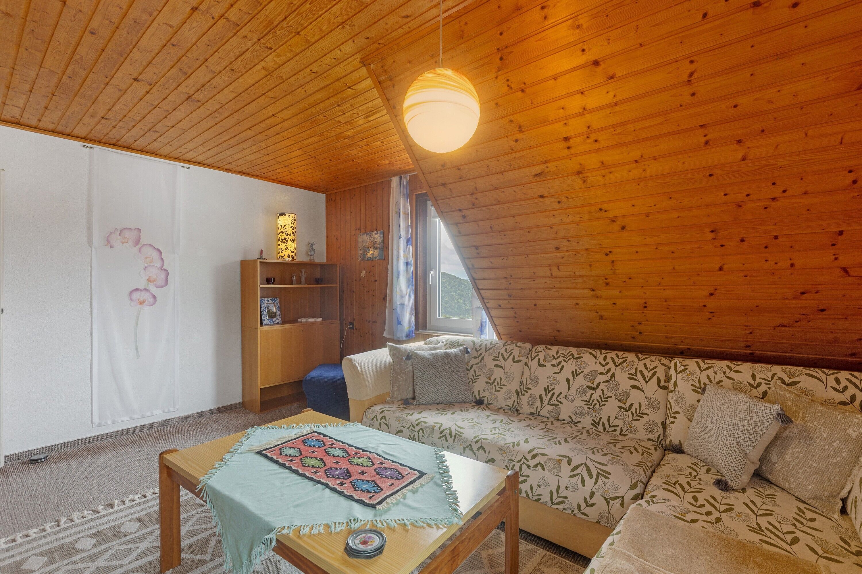 Una stanza accogliente con soffitto in legno, un divano rivestito di fiori, un tavolino con tovaglia e una libreria.