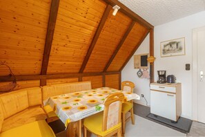 Une pièce chaleureuse avec un plafond en bois, une table recouverte d’une nappe à motifs de tournesols, des chaises jaunes, une petite kitchenette et un tableau encadré accroché au mur.