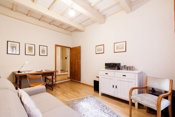 A living room with a sofa, a dining table with chairs, a cabinet with books, and framed pictures on the wall.