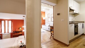 A room with a bed, a desk, a chair, a window, and a view of a kitchen area.