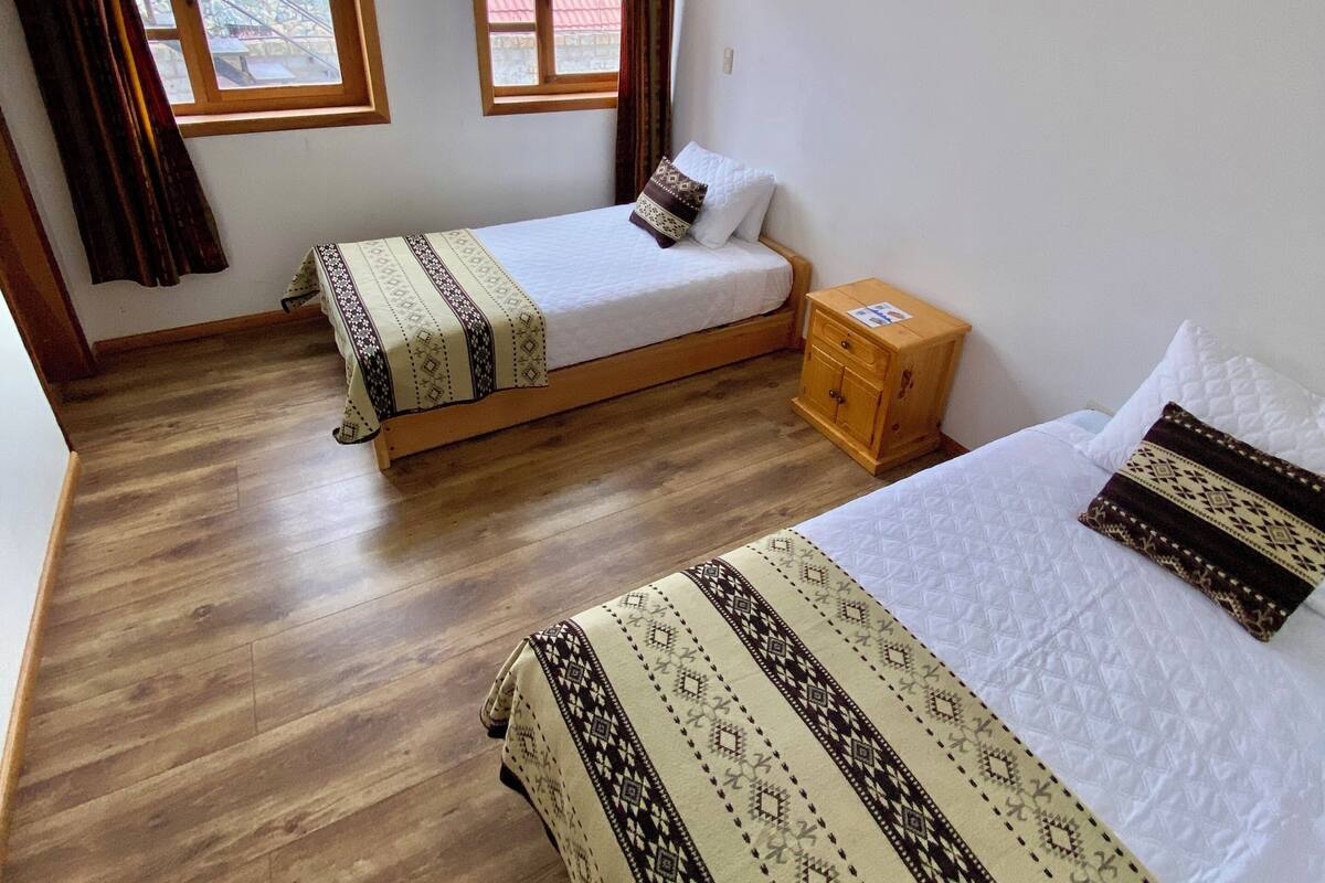A hotel room with two beds, wooden flooring, a window with curtains, and a nightstand.