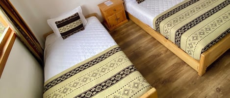 Two single beds with patterned bedding, a wooden nightstand, and a small wooden cabinet in a room with wooden flooring and white walls.