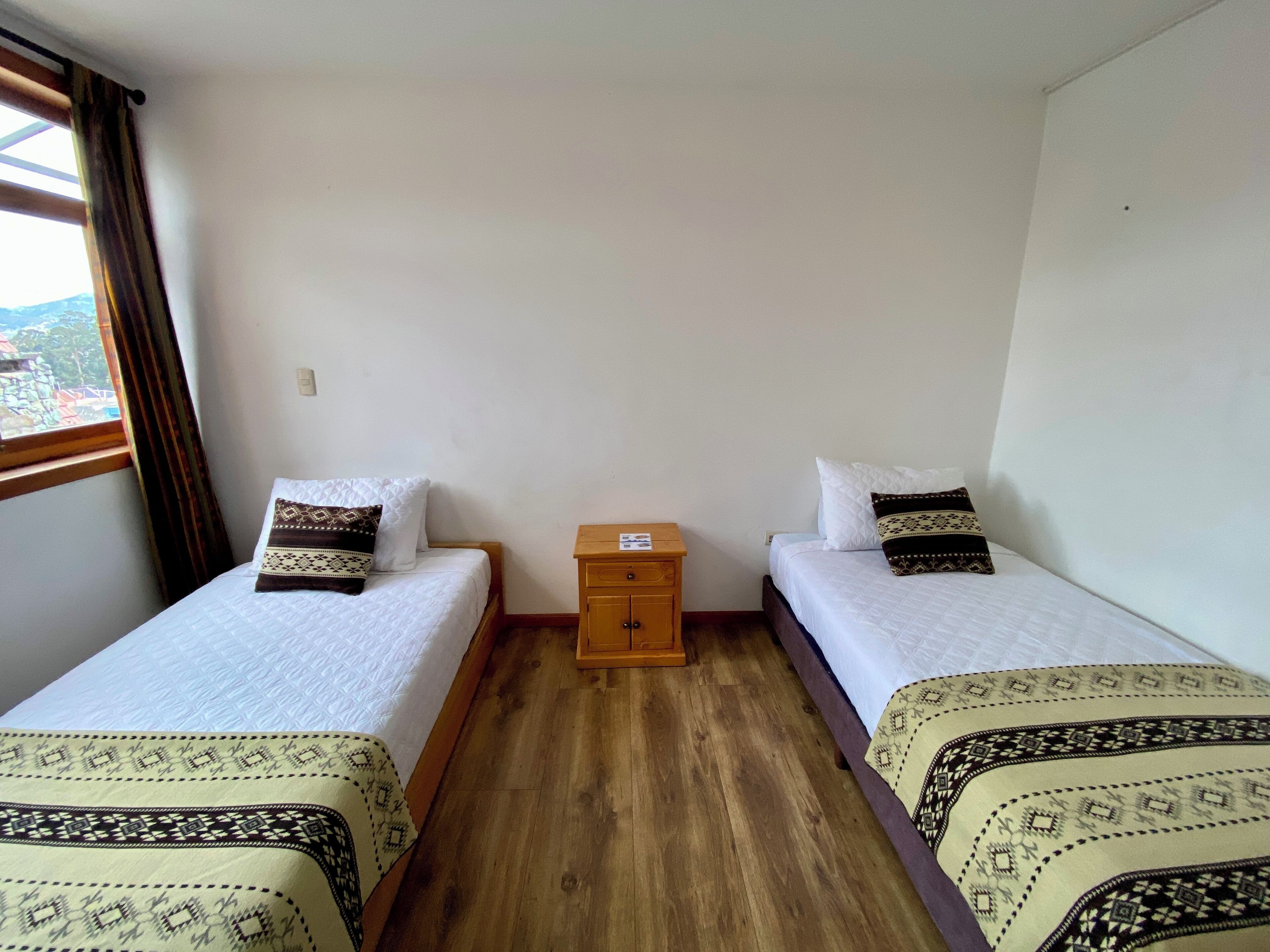 A room with two single beds, a small wooden nightstand, and a window with curtains.