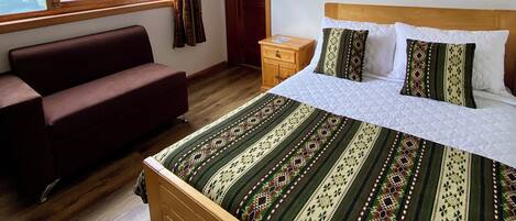 A bedroom with a bed, a wooden headboard, a wooden nightstand, a brown sofa, a window with curtains, and a wooden floor.