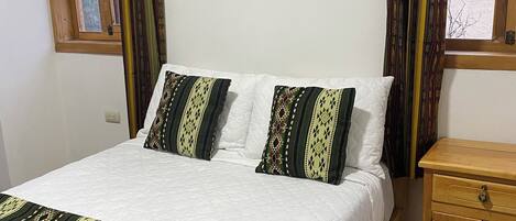A bedroom with a bed, patterned pillows, a wooden nightstand, and curtains.