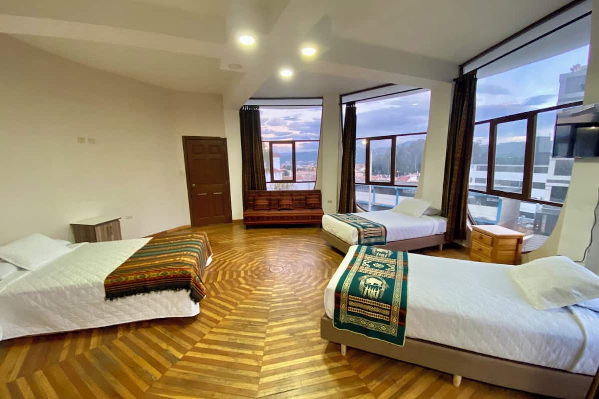 A hotel room with two beds, a sofa, a wooden floor, and large windows offering a city view.