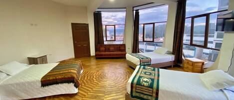 A hotel room with two beds, a sofa, a wooden floor, and large windows offering a city view.