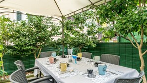 Ett utomhusområde med plats för måltider, där ett bord är dukat för fyra personer, omgivet av flätade stolar, under ett tak.