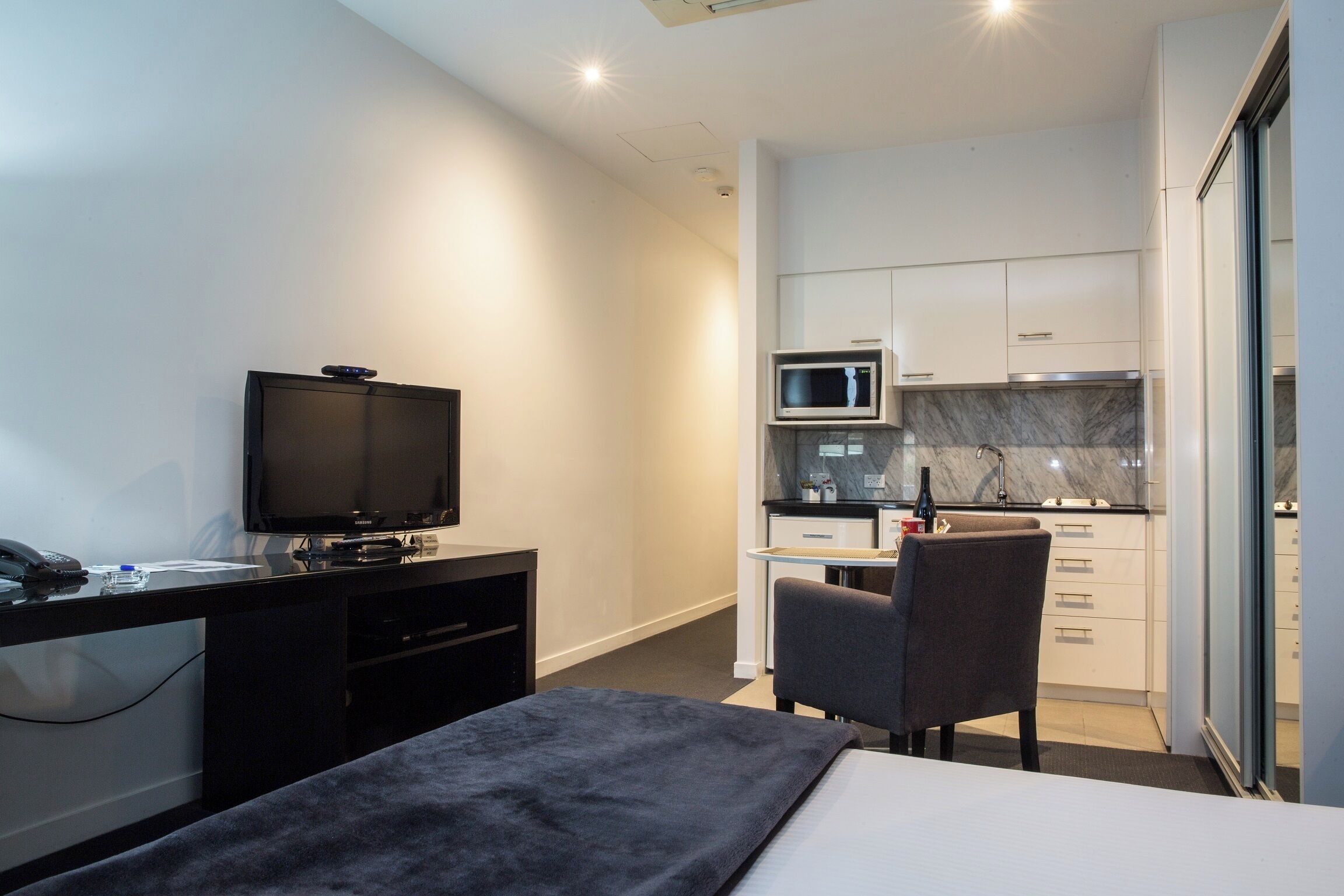 A compact hotel room with a bed, a desk with a television, a small kitchenette, and a chair.
