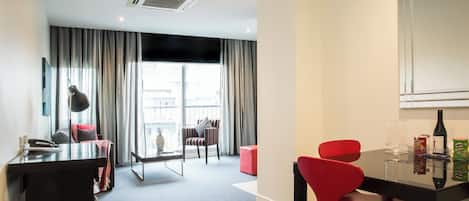 Una habitación de hotel moderna con escritorio, sillas rojas, una mesa de comedor y un amplio ventanal con cortinas.