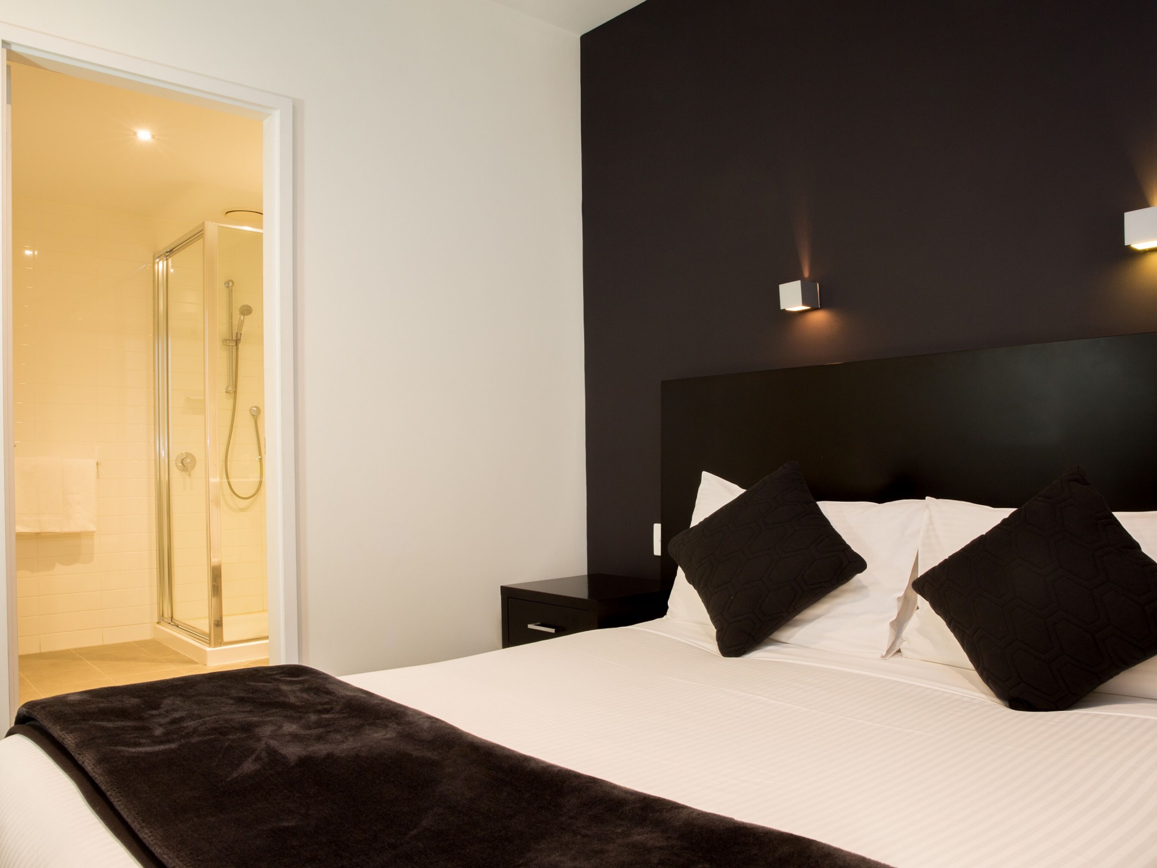 Ein modernes Hotelzimmer mit einem großen Bett, einem dunklen Kopfteil und einer Duschkabine aus Glas.