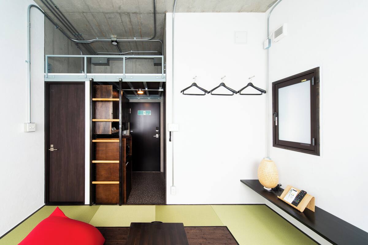A room with a loft bed, a wooden wardrobe, a shelf with a lamp, and a wall-mounted mirror.