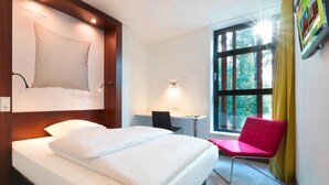 Ein Hotelzimmer mit Bett, Schreibtisch, Stuhl, einem Fenster mit Vorhängen und einem an der Wand befestigten Fernseher.