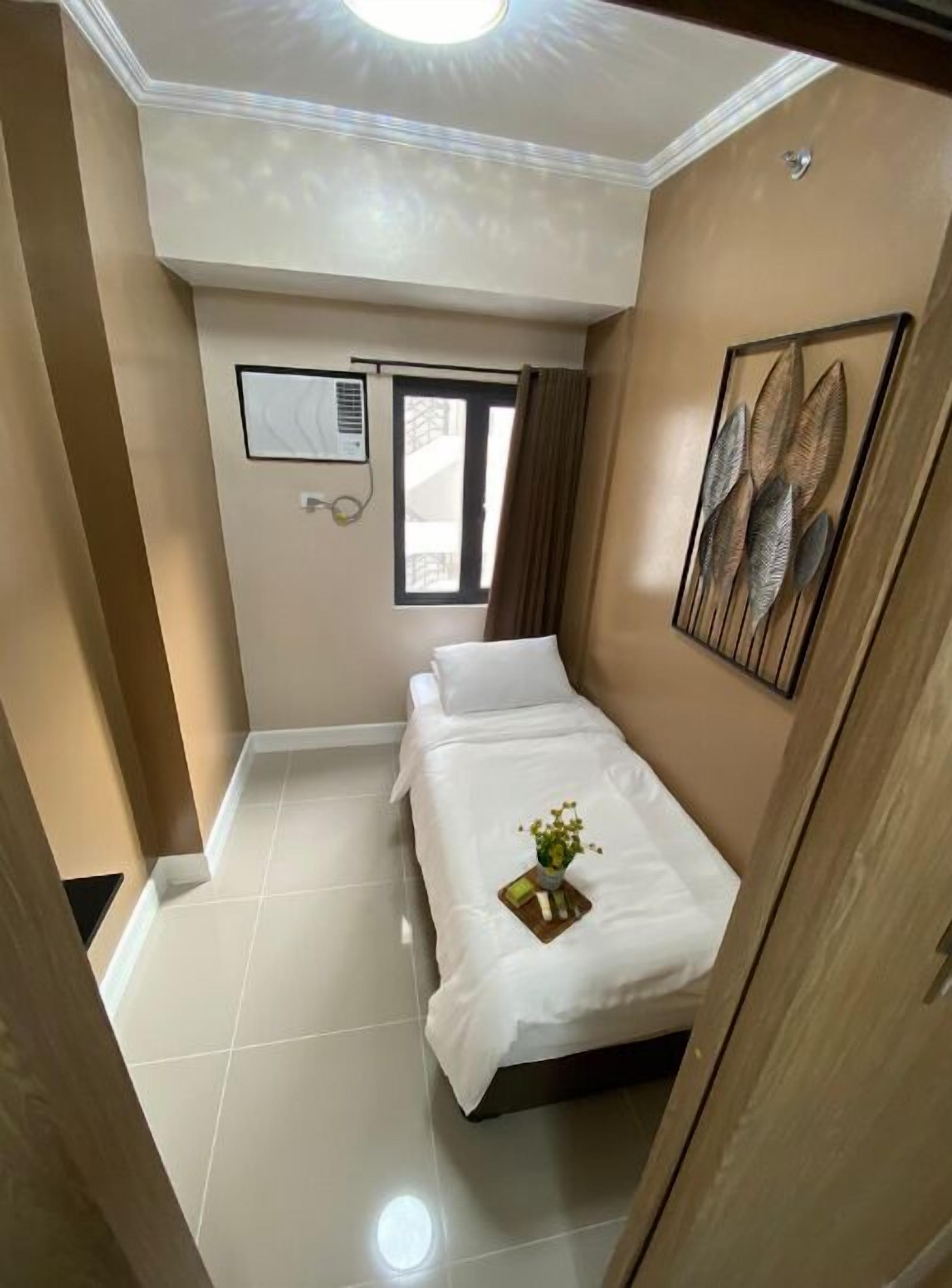 A small, well-lit bedroom with a bed, a window, and a wall-mounted air conditioning unit.