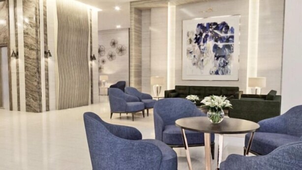 Een moderne lobby met een marmeren receptiebalie, blauwe gestoffeerde fauteuils, een ronde tafel met een vaas en een plafondventilator.
