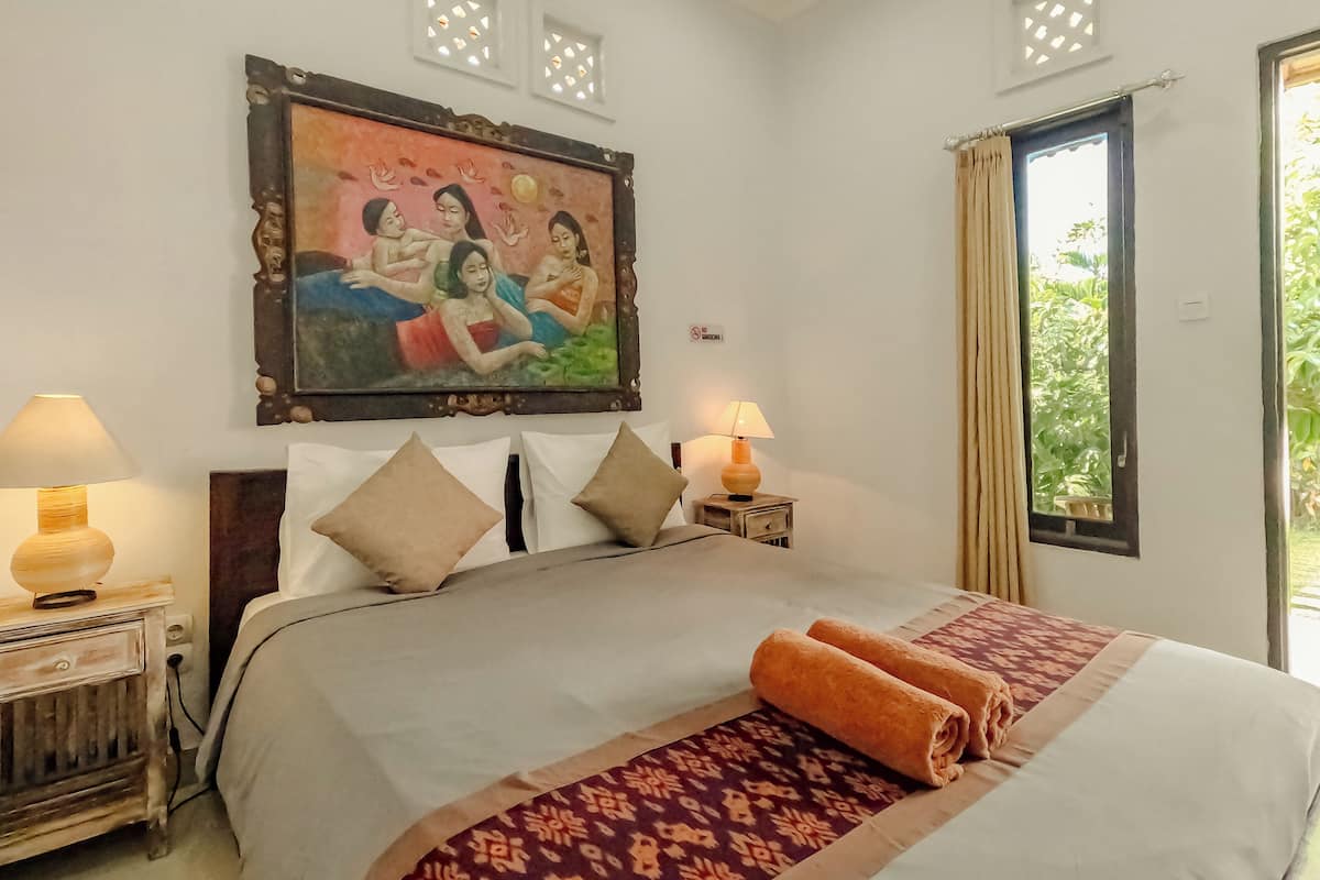 A neatly made bed with a patterned bedspread, two bedside lamps, a painting above the bed, and a window with curtains.