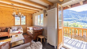 A cozy log cabin interior with a wooden ceiling, a sofa, a brown leather couch, a small wooden table, and a view of a mountainous landscape through a balcony.