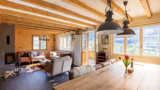 A spacious living area with a fireplace, wooden beams, a large dining table, and a view of the outdoors.