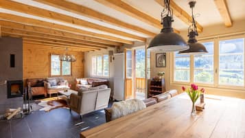 Een ruime woonkamer met een open haard, houten balken, een grote eettafel en uitzicht op de omgeving.