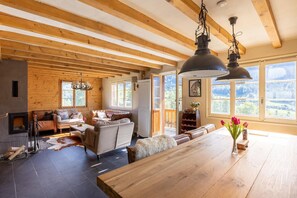 En rummelig opholdsstue med pejs, træbjælker, et stort spisebord og udsigt til det fri.