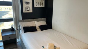 A neatly made bed with white linens and black pillows, a bedside table with a lamp, and a window with a view of buildings. - 2 Bed with Wi‑Fi, Near Central Birmingham (Birmingham)