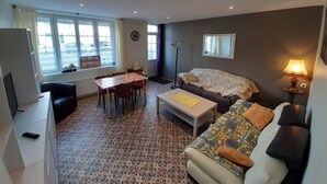 A furnished living room with a sofa, a coffee table, a dining table with chairs, a TV, and a window with a view of the street.