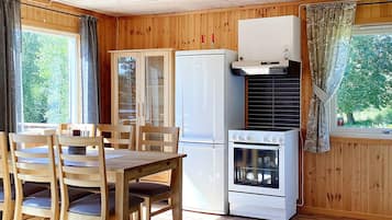 A compact kitchen with wooden walls, a dining table with chairs, a refrigerator, an oven, and a window with curtains.