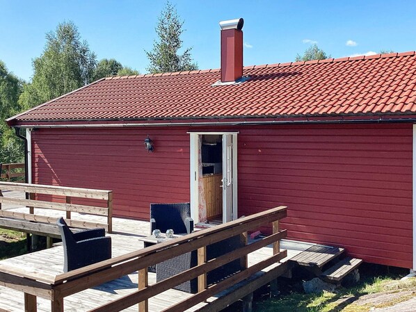 A red wooden cabin with a red-tiled roof, a chimney, and a wooden deck with a chair and table.