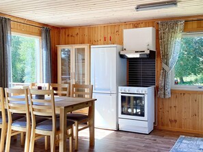 A compact kitchen with wooden walls, a dining table with chairs, a refrigerator, an oven, and a window with curtains.