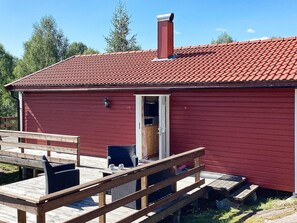 A red wooden cabin with a red-tiled roof, a chimney, and a wooden deck with a chair and table. - 4 Person Holiday Home in Bengtsfors (Bengtsfors)